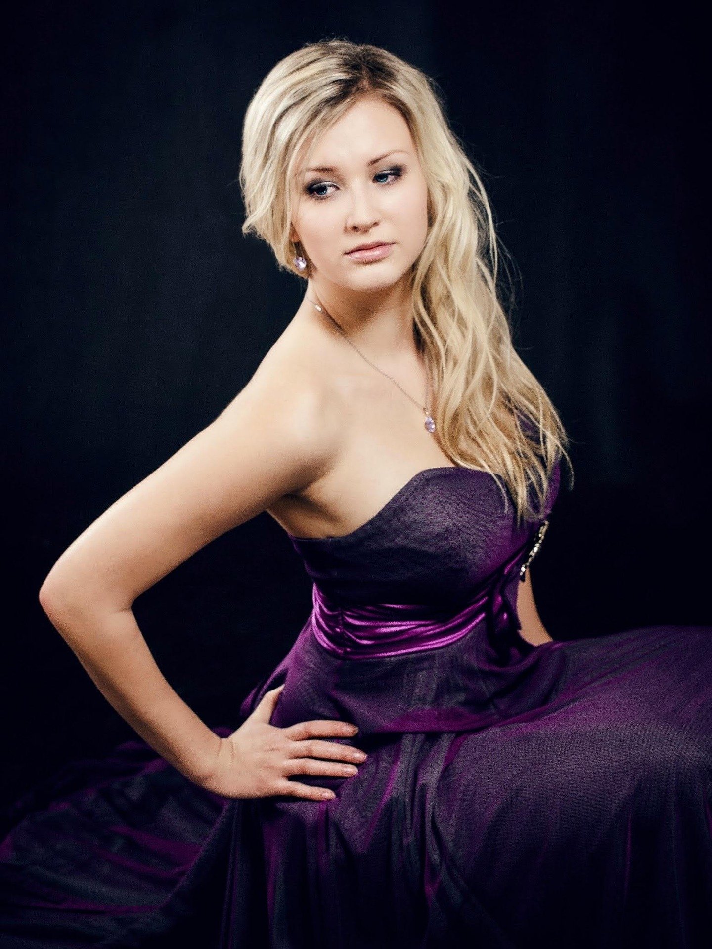 Woman posing in a strapless purple satin evening gown against a dark background